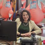 A woman sits at her desk, smiling, with inflatable flamingos behind her. Signs reading 'You've been flocked' are placed among the decorations