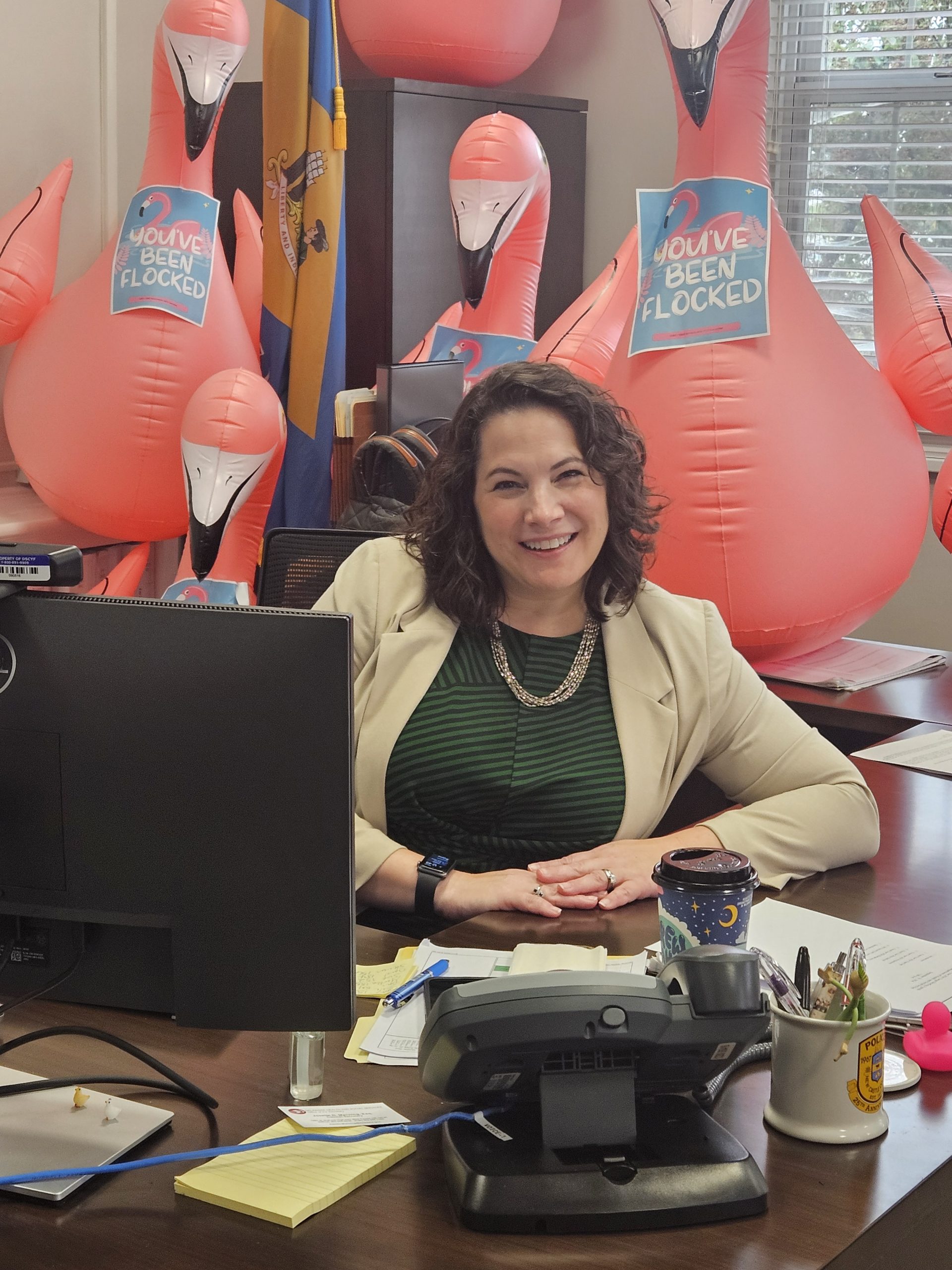 A woman sits at her desk, smiling, with inflatable flamingos behind her. Signs reading 'You've been flocked' are placed among the decorations