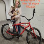 A woman smiles while holding her dog, the first-place winner of the Cutest Pet Photo Contest. Next to her is the prize, a bright red bike.