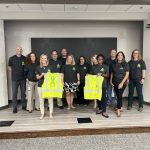 DelDOT Leadership Team members wear mental health awareness t-shirts, with two members holding safety vests—one displaying the front and the other showing the back—during a fundraiser for ContactLifeLine.