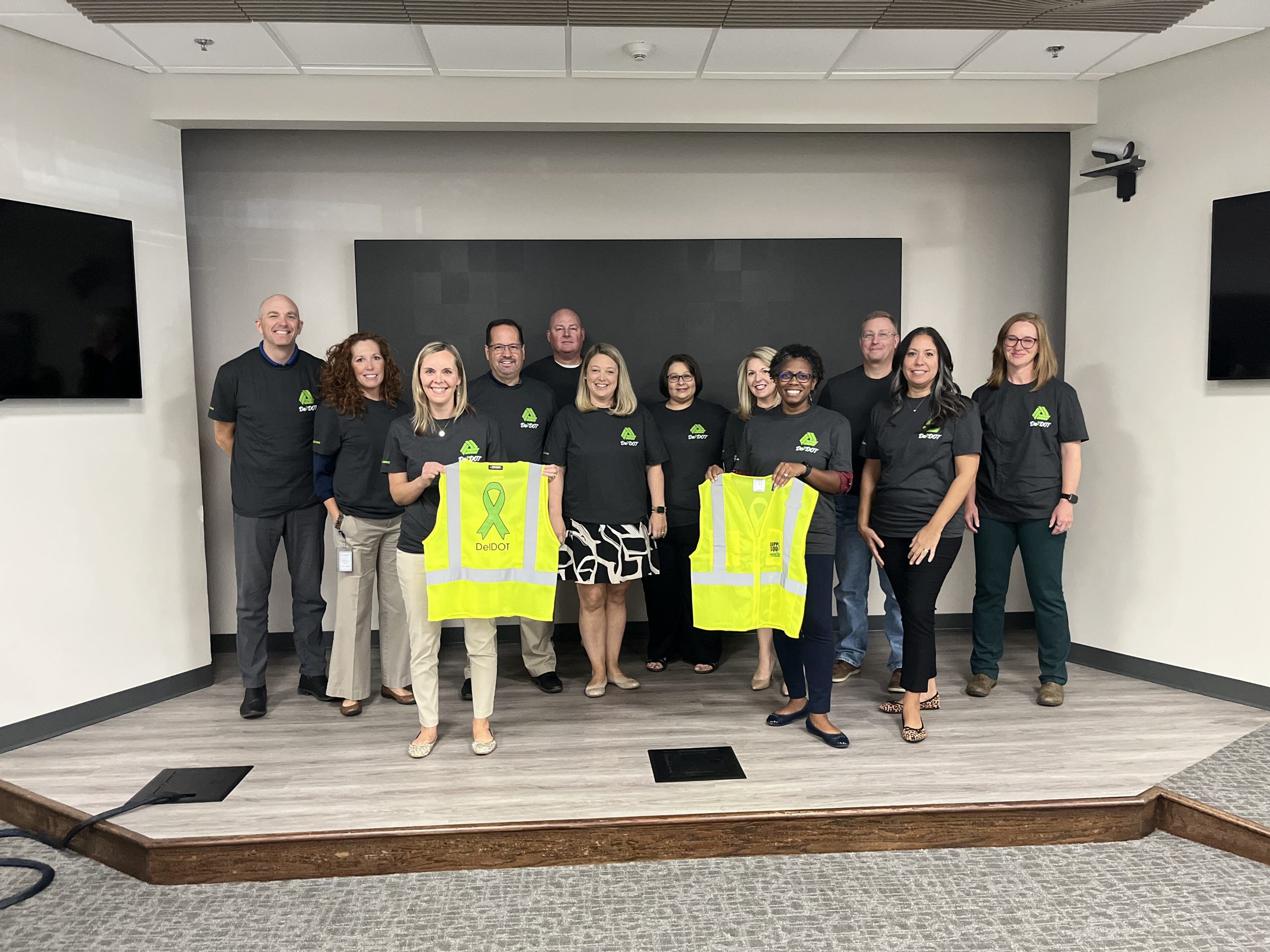 DelDOT Leadership Team members wear mental health awareness t-shirts, with two members holding safety vests—one displaying the front and the other showing the back—during a fundraiser for ContactLifeLine.