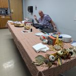 A woman waves to the camera behind a table decorated with fall-themed decorations and grilled cheese supplies, as part of the STEMS team’s fundraiser for the State Employees' Charitable Campaign.