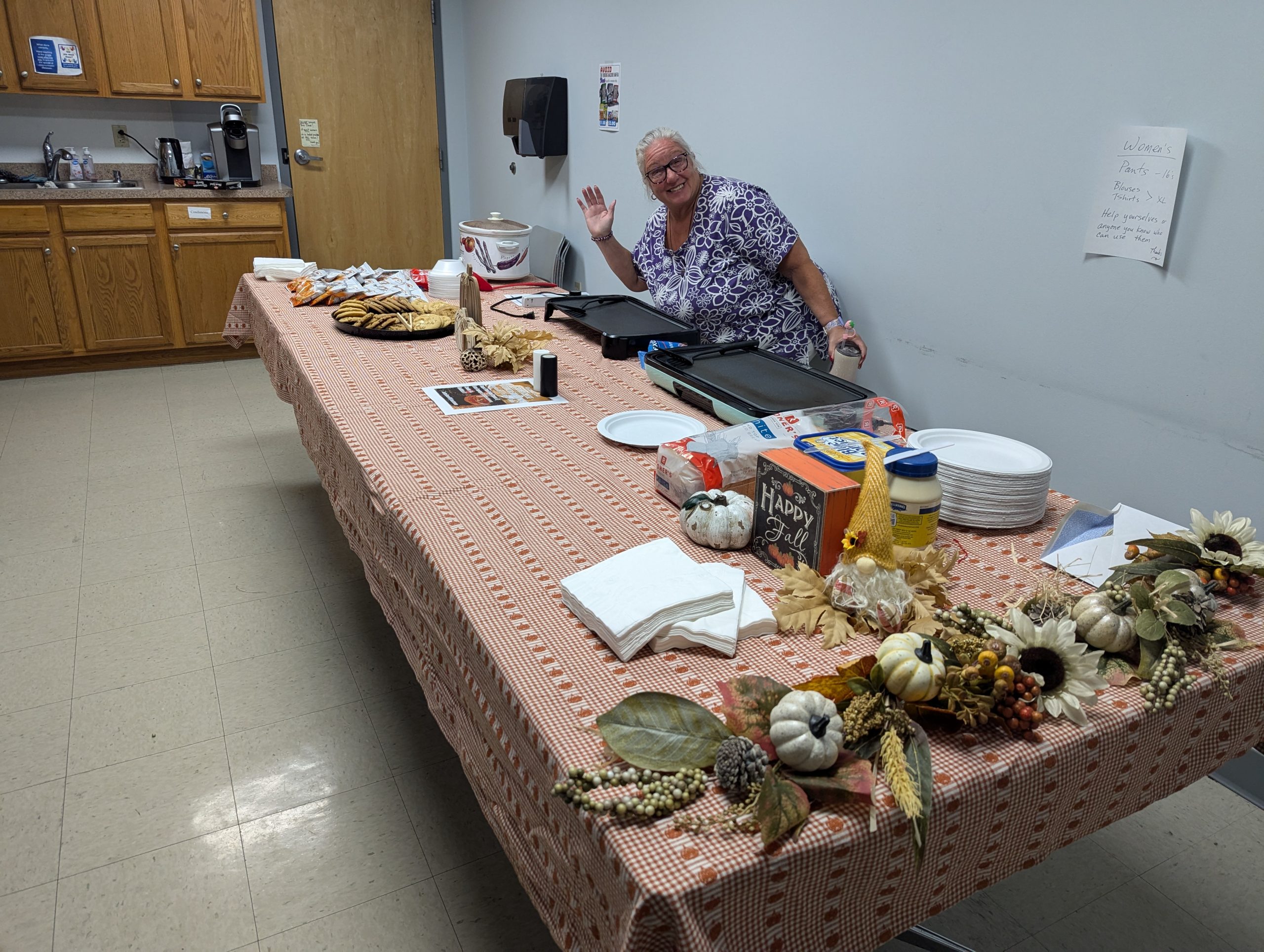 A woman waves to the camera behind a table decorated with fall-themed decorations and grilled cheese supplies, as part of the STEMS team’s fundraiser for the State Employees' Charitable Campaign.